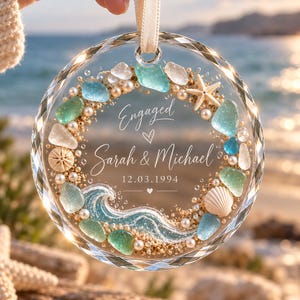 K&ouml;nnte beinhalten: Ein rundes, facettiertes Glasornament mit einem Kranzdesign im Strandthema. Es enth&auml;lt Meerglas, kleine Perlen, Sand, einen Seestern und eine Muschel. Der Text lautet Engaged, Sarah & Michael und 12.03.1994, mit einer blauen Welle.