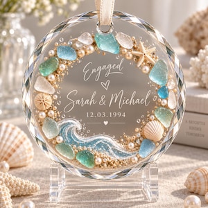 K&ouml;nnte beinhalten: Ein kreisf&ouml;rmiges Glasornament mit Strandmotiv, verziert mit Meerglas, kleinen Perlen, einem Seestern und einer Muschel. In der Mitte steht der Text Engaged, Sarah & Michael sowie das Datum 12.03.1994, erg&auml;nzt durch eine gemalte blaue Meereswelle.