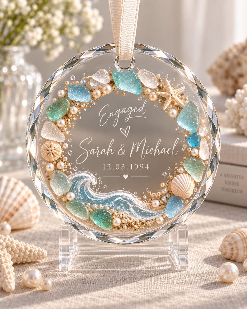 K&ouml;nnte beinhalten: Ein kreisf&ouml;rmiges Glasornament mit Strandmotiv, verziert mit Meerglas, kleinen Perlen, einem Seestern und einer Muschel. In der Mitte steht der Text Engaged, Sarah & Michael sowie das Datum 12.03.1994, erg&auml;nzt durch eine gemalte blaue Meereswelle.