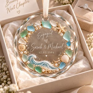 K&ouml;nnte beinhalten: Ein kreisf&ouml;rmiges Glasornament mit facettiertem Rand, verziert mit Meerglas, Perlen, einem Seestern und einer Muschel. Der Text lautet Engaged, Sarah & Michael und 12.03.1994. Es h&auml;ngt an einem cremefarbenen Band in einer Geschenkbox.