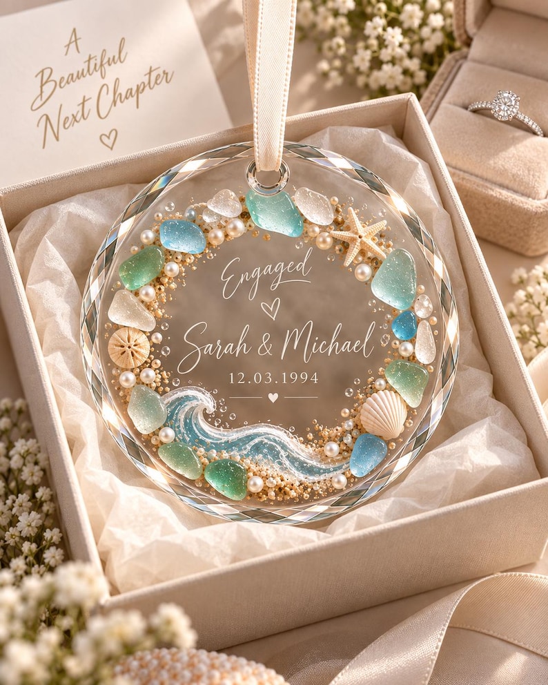 K&ouml;nnte beinhalten: Ein kreisf&ouml;rmiges Glasornament mit facettiertem Rand, verziert mit Meerglas, Perlen, einem Seestern und einer Muschel. Der Text lautet Engaged, Sarah & Michael und 12.03.1994. Es h&auml;ngt an einem cremefarbenen Band in einer Geschenkbox.