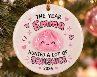 Könnte beinhalten: Ein runder weißer Keramikschmuck mit einer goldenen Aufhängeschnur. Der Text lautet THE YEAR Emma HUNTED A LOT OF SQUISHIES 2026. In der Mitte befindet sich eine rosa glitzernde Knödel-Figur mit lächelndem Gesicht, umgeben von kleinen rosa Herzen.