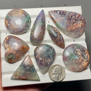 May include: Eight polished cabochons in various shapes including circles, hearts, and triangles, featuring a translucent base with purple, green, and copper-colored metallic inclusions. A US nickel is included for scale next to the numbered stones.
