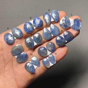 May include: Eleven pairs of blue sapphire gemstones in diverse shapes, including oval and teardrop. Each stone is faceted, showing a range of blue hues and internal patterns. The gemstones are displayed on white paper backing.