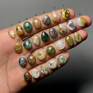 Puede incluir: Una colección de treinta piedras preciosas pulidas de diversas formas y colores, incluyendo verde, marrón, amarillo y blanco. Cada piedra está numerada del 1 al 30, mostrando una variedad de patrones y texturas naturales. Las piedras preciosas se sostienen en una mano.