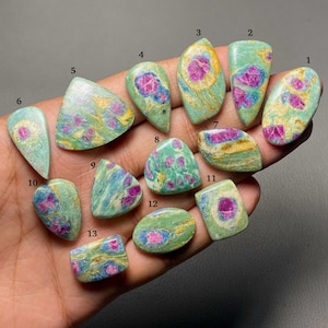 May include: Thirteen polished cabochon gemstones labeled one through thirteen, featuring a mix of vibrant pink ruby inclusions embedded within a light green and blue fuchsite matrix, displaying varied shapes including oval, teardrop, and rectangular.