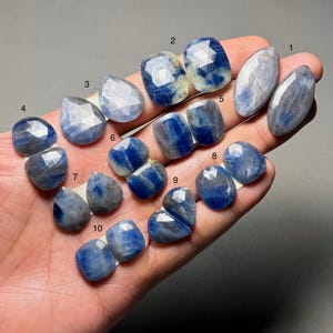 May include: A selection of ten faceted blue sapphire gemstones in diverse shapes, such as oval, teardrop, and cushion cuts. The sapphires exhibit a spectrum of blue shades, from light to deep indigo, with some stones showing a mix of colours.