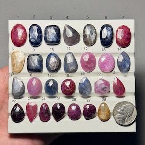 Op de afbeelding: Een verzameling gefacetteerde edelstenen in diverse vormen en kleuren, waaronder roze, blauw en paars. De edelstenen zijn gerangschikt op een wit displaybord, met een munt voor schaal. De edelstenen zijn genummerd van 1 tot en met 28. Afmetingen in centimeters.