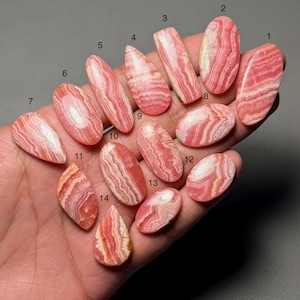 Puede incluir: Una colección de catorce piedras preciosas de rodocrosita pulidas en varias formas, incluyendo ovaladas, en forma de lágrima y rectangulares. Las piedras muestran una mezcla de patrones de bandas rosas y blancas, algunas con un tono rojo más intenso. Las piedras preciosas se sostienen en una mano.