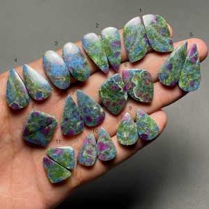 Puede incluir: Una colección de once pares de piedras preciosas de rubí en fuchsite pulidas, con formas de lágrima y escudo. Cada piedra muestra un patrón moteado de inclusiones minerales en magenta vibrante, verde brillante y azul cielo, numeradas del uno al once.
