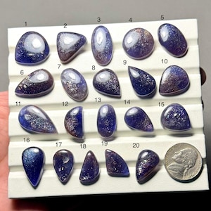 Puede incluir: Una colección de veinte piedras preciosas pulidas, de diversas formas, incluyendo óvalos y lágrimas. Las piedras muestran una mezcla de tonos azul profundo, púrpura y plateado, con una apariencia brillante y moteada. Se incluye una moneda como referencia de tamaño.