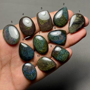 Puede incluir: Una colección de once piedras preciosas de obsidiana pulidas en varias formas, incluyendo ovaladas, en forma de lágrima y rectangulares. Las piedras muestran colores iridiscentes de verde, azul y dorado, con un acabado brillante. Las piedras preciosas se sostienen en una mano.