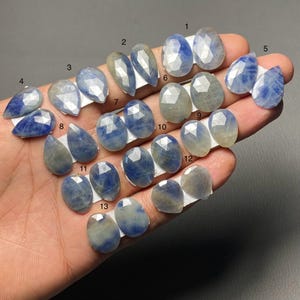 Puede incluir: Una colección de piedras preciosas de zafiro azul facetadas en varias formas, incluyendo ovaladas, pera y lágrima. Las piedras se exhiben en pequeñas tarjetas blancas, mostrando su color y claridad. Los zafiros varían en color de azul claro a oscuro.