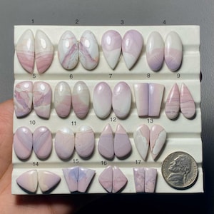 May include: A collection of seventeen polished gemstones in various shapes and shades of pink, lavender, and white, displayed on a white tray. The gemstones include oval, teardrop, and geometric cuts. A US nickel is included for size reference.