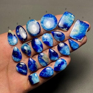 May include: A collection of twenty polished gemstones in various shapes, including oval, teardrop, and rectangular. The stones display a mix of deep blue and white hues, with some featuring a marbled effect. The gemstones are displayed against a neutral background.