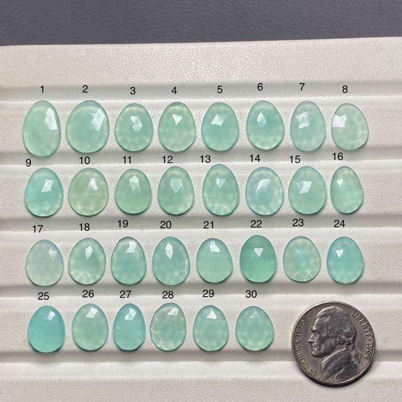 Peut inclure: Une collection de 30 cabochons de pierres pr&eacute;cieuses bleu clair, chacun avec une forme et une taille uniques, dispos&eacute;s en rang&eacute;es. Une pi&egrave;ce de monnaie est incluse pour la comparaison des tailles. Les pierres pr&eacute;cieuses varient en clart&eacute; et en facettage.