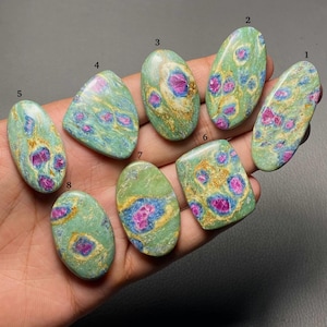 May include: Eight polished cabochon gemstones labeled one through eight, featuring a light green matrix with embedded vibrant pink and blue mineral inclusions. Each stone has a smooth, rounded surface with distinct color patterns and natural textures.
