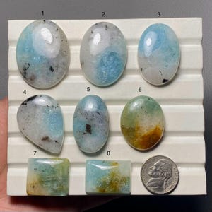 Può includere: Una collezione di otto pietre preziose lucide di varie forme, tra cui ovali e rettangoli, esposte su un vassoio bianco. Le pietre presentano un mix di colori, tra cui azzurro, bianco e marrone, con una moneta da cinque centesimi americana per il confronto delle dimensioni.