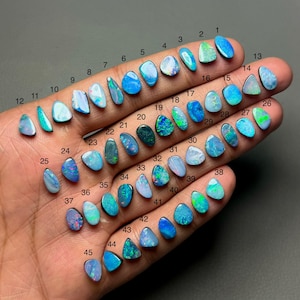 May include: A hand holds 45 opals, each with a distinct shape and colour. The opals vary from light blue to dark green, with flashes of other colours. Each opal is numbered for identification.