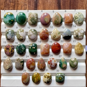 Puede incluir: Una colección de treinta y dos piedras preciosas cabujón pulidas dispuestas en una bandeja de exhibición blanca. Cada piedra presenta patrones únicos, incluyendo orbes circulares, texturas moteadas y colores variados como verde, rojo.