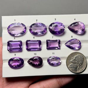 Könnte beinhalten: Eine Auswahl von elf Amethyst-Edelsteinen in verschiedenen Formen, darunter ovale, birnenförmige und rechteckige Schliffe. Die Edelsteine variieren in der Farbe von hell- bis tiefviolett, mit einer Münze zum Größenvergleich.