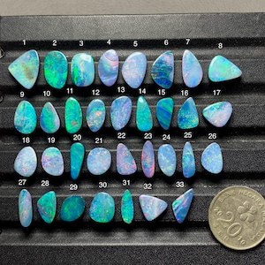 May include: A collection of thirty-two polished, multi-colored opals arranged in rows on a black display tray, each numbered from 1 to 33, excluding 27, with a 2010 Bank Negara 20 sen coin included for scale, showing vibrant blue and green flashes.