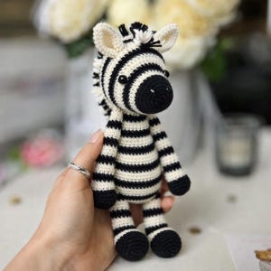 May include: A handmade crochet zebra toy with black and white stripes. The zebra has a black snout, ears, and hooves. The toy is held in a person's hand, showcasing the detailed craftsmanship of the stuffed animal. The background is blurred, with flowers and a candle holder.