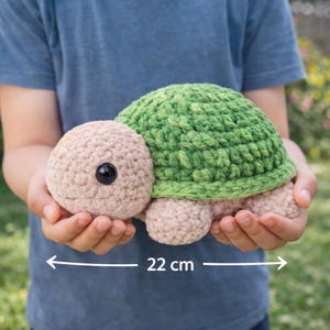 May include: A handmade crochet turtle toy, featuring a green shell, a tan head, and tan feet. The turtle has a black eye and measures 22 cm (8.6 inches) in length. The toy is held in a person's hands.