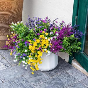May include: A white, ribbed ceramic planter overflowing with colorful faux flowers. The arrangement includes yellow, purple, white, and green foliage, creating a vibrant display. The planter sits on a gray stone surface, near a green door.