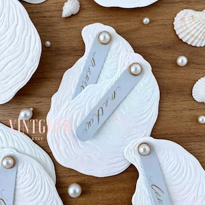 Puede incluir: Tarjetas de mesa blancas en forma de concha con una superficie texturizada, con un acento de perla y una etiqueta con el nombre. Las etiquetas con el nombre son de color azul claro con letras doradas. Perlas y conchas marinas dispersas completan el tema costero.