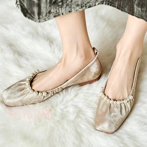 May include: A pair of champagne-colored velvet ballet flats. The square-toe shoes are adorned with a row of small pearls along the top edge. The soles are a light brown color. The shoes are on a white, fluffy surface.