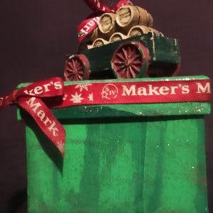 May include: A green box-shaped ornament with a miniature wagon carrying small wooden barrels. The wagon has red wheels. A red ribbon with the text "Maker's Mark" is tied around the box and wagon.