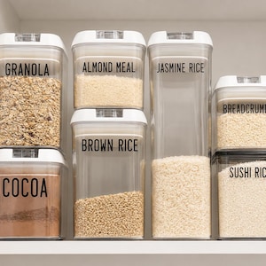 May include: A collection of clear rectangular plastic food storage containers with white lids, organized on a shelf. The containers are labeled with black text reading Granola, Almond Meal, Jasmine Rice, Breadcrumbs, Brown Rice, Cocoa, and Sushi Rice.