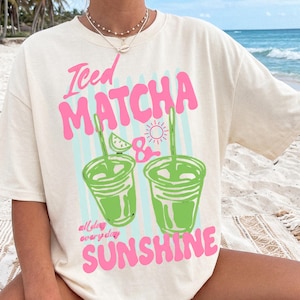 May include: A cream-colored short-sleeve t-shirt featuring a graphic print with the text Iced Matcha & all day everyday SUNSHINE. The design includes two green iced matcha drinks with straws, a lime wedge, and a small pink sun icon.