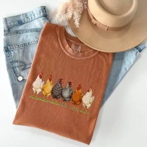May include: A rust-colored t-shirt featuring a graphic of six chickens in various colors standing in a row on a green patch. The shirt is paired with light blue denim jeans, a tan wide-brimmed hat, and a silver chain necklace with a small pendant.