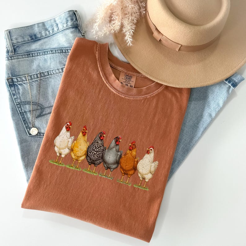 May include: A rust-colored t-shirt featuring a graphic of six chickens in various colors standing in a row on a green patch. The shirt is paired with light blue denim jeans, a tan wide-brimmed hat, and a silver chain necklace with a small pendant.