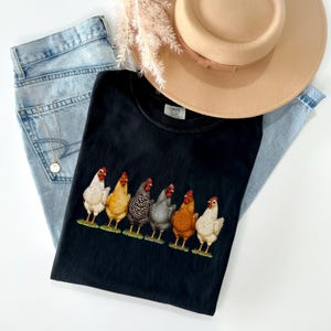 May include: A black t-shirt featuring a graphic print of six chickens in various colors including white, yellow, barred, grey, and brown, standing in a row. The shirt is paired with light blue denim jeans, a tan wide-brimmed hat, and a gold necklace.