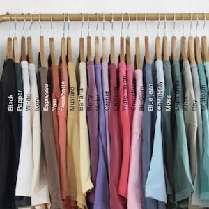May include: A collection of t-shirts hanging on a gold metal rack, displayed in a gradient of colors labeled Black, Pepper, White, Ivory, Espresso, Yam, Terracotta, Mustard, Banana, Berry, Violet, Orchid, Crimson, Watermelon, Crunchberry, Blossom, Blue jean, Chambray, Moss, Light Green, Khaki, Bay, Seafoam, Island Reef, and Chalky Mint.