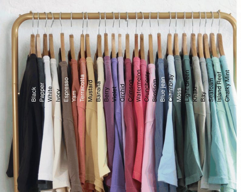 May include: A collection of t-shirts hanging on a gold metal rack, displayed in a gradient of colors labeled Black, Pepper, White, Ivory, Espresso, Yam, Terracotta, Mustard, Banana, Berry, Violet, Orchid, Crimson, Watermelon, Crunchberry, Blossom, Blue jean, Chambray, Moss, Light Green, Khaki, Bay, Seafoam, Island Reef, and Chalky Mint.