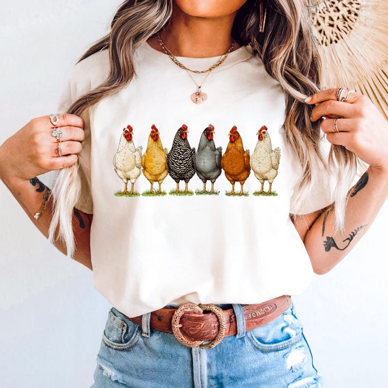 May include: A cream-colored short-sleeve t-shirt featuring a graphic print of six chickens standing in a row. The chickens are depicted in various colors including white, yellow, black and white barred, grey, brown, and light tan, all on green grass.