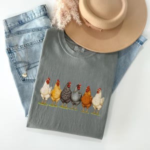 May include: A charcoal gray crew neck t-shirt featuring a graphic print of six chickens in various colors including white, yellow, black and white striped, gray, and brown, standing in a row on a thin strip of green grass.