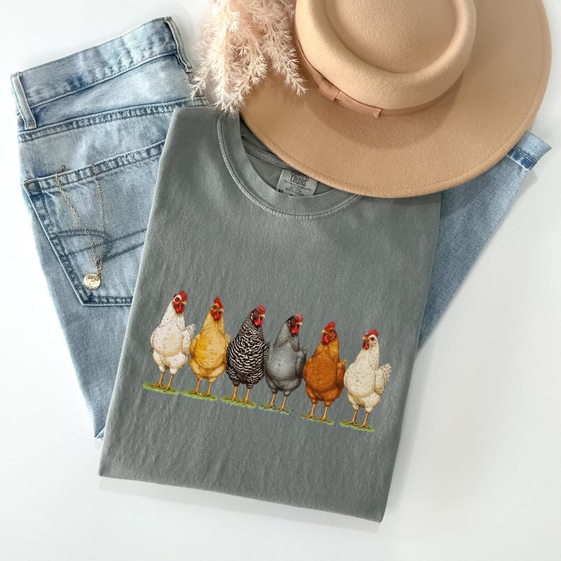May include: A charcoal gray crew neck t-shirt featuring a graphic print of six chickens in various colors including white, yellow, black and white striped, gray, and brown, standing in a row on a thin strip of green grass.