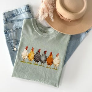May include: A sage green t-shirt featuring a graphic print of six chickens in various colors including white, yellow, black and white striped, dark gray, and brown. The shirt is displayed with light blue denim jeans, a tan fedora, and a silver necklace.