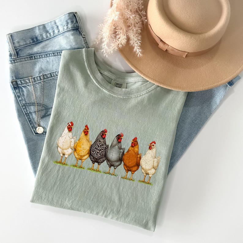 May include: A sage green t-shirt featuring a graphic print of six chickens in various colors including white, yellow, black and white striped, dark gray, and brown. The shirt is displayed with light blue denim jeans, a tan fedora, and a silver necklace.