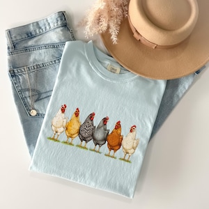 May include: A light blue t-shirt featuring a graphic of six chickens in various colors including white, yellow, black and white striped, grey, and brown. The shirt is paired with light wash blue denim jeans, a tan wide-brimmed hat, and a silver necklace.