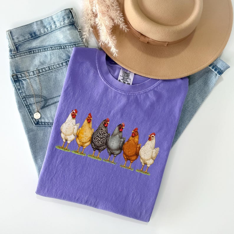 May include: A purple t-shirt featuring a graphic print of six chickens in various colors including white, yellow, black and white striped, grey, and brown, standing on a green patch. The shirt has a crew neckline and a Comfort Colors brand label.