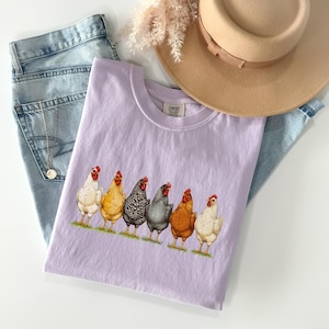 May include: A light purple t-shirt featuring a graphic of six chickens in various colors standing in a row. The shirt is paired with light blue denim jeans, a tan wide-brimmed hat, and a silver necklace with a small circular pendant.