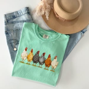 May include: A mint green t-shirt featuring a graphic of six chickens in various colors, including white, yellow, black and white speckled, grey, and brown. The shirt is paired with light blue denim jeans and a tan wide-brimmed felt hat.