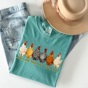 May include: A teal short-sleeved t-shirt featuring a graphic print of six chickens in various colors including white, yellow, black and white striped, grey, and brown. The shirt has a crew neckline and a Comfort Colors brand label at the neck.