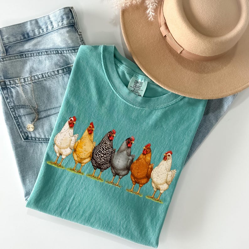 May include: A teal short-sleeved t-shirt featuring a graphic print of six chickens in various colors including white, yellow, black and white striped, grey, and brown. The shirt has a crew neckline and a Comfort Colors brand label at the neck.
